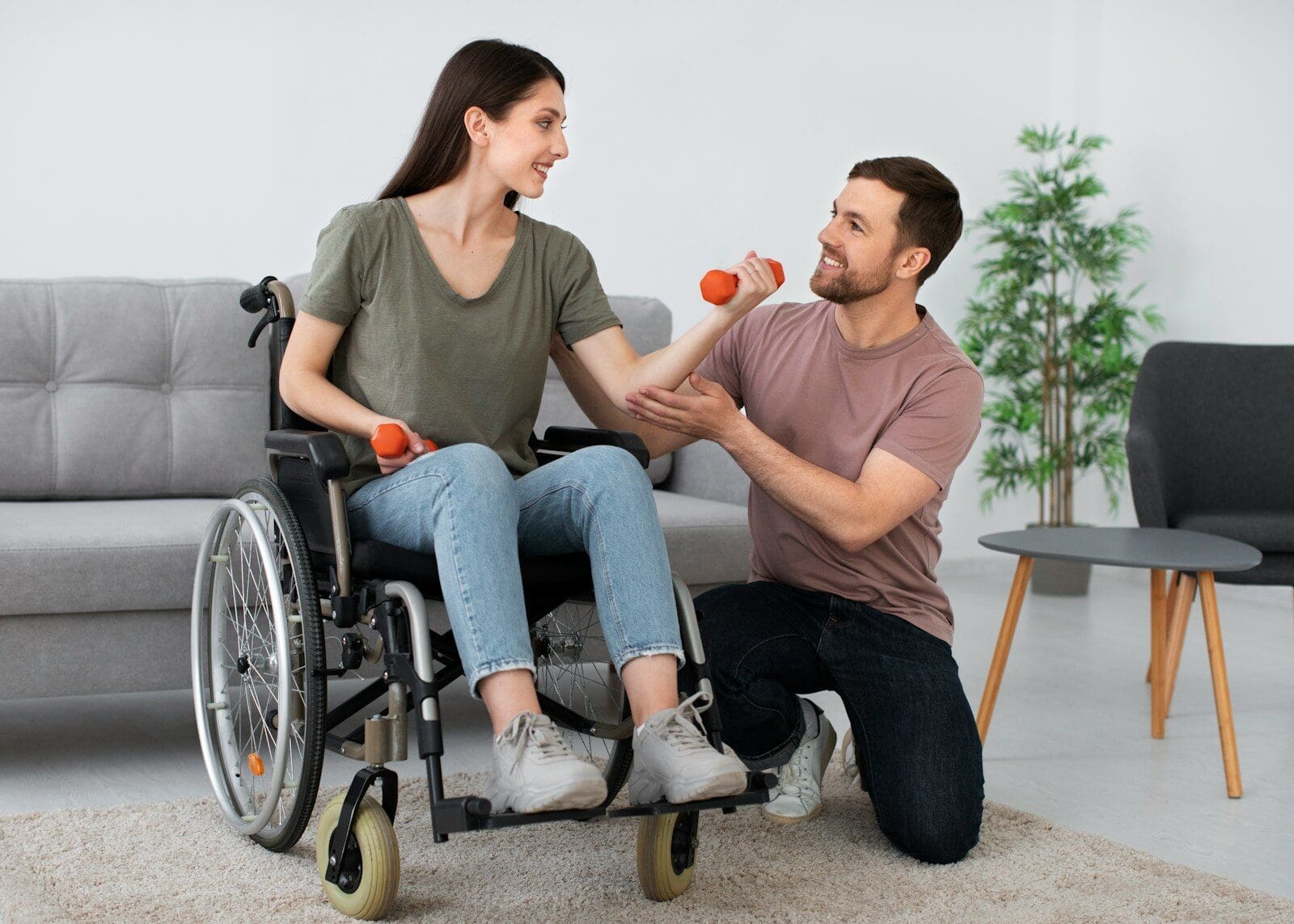 A woman in a wheelchair lifting orange dumbbells with a man kneeling beside her